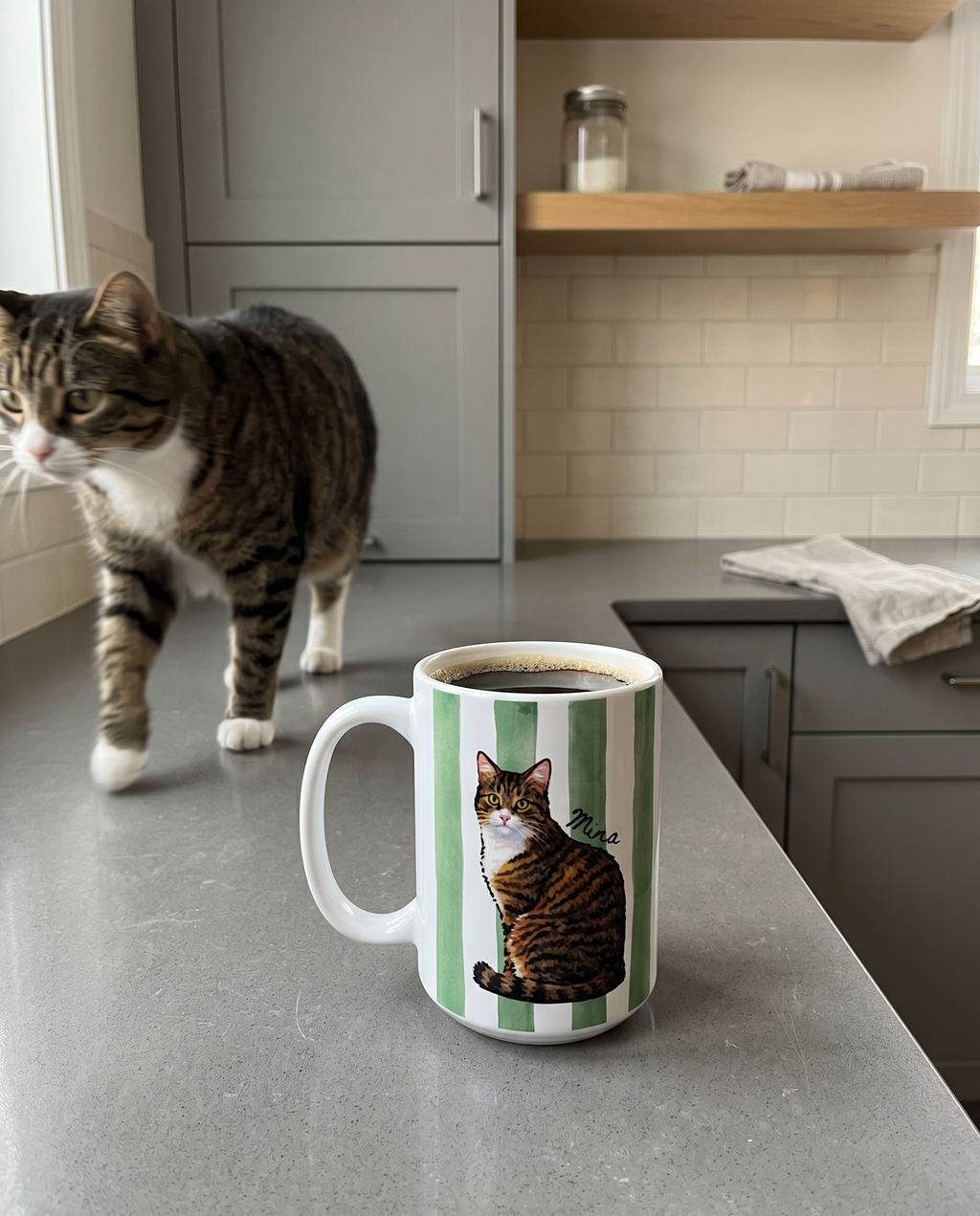 A white ceramic mug with green vertical stripes and an illustration of a tabby cat, filled with coffee and placed on a gray kitchen counter, with a real tabby cat walking in the background.