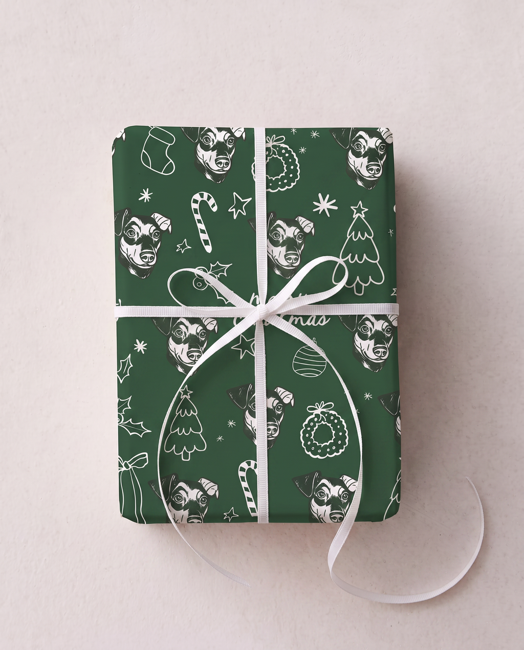A rectangular gift box wrapped in dark green Christmas-themed paper with white line-art illustrations of stockings, candy canes, ornaments, and trees, tied with a white ribbon bow on a light beige background.