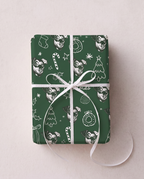 A rectangular gift box wrapped in dark green Christmas-themed paper with white line-art illustrations of stockings, candy canes, ornaments, and trees, tied with a white ribbon bow on a light beige background.