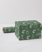 Green Christmas-themed wrapping paper with white illustrations of dog faces, candy canes, ornaments, and 'merry christmas' text, shown wrapped around a box with a matching roll beside it on a light gray background.