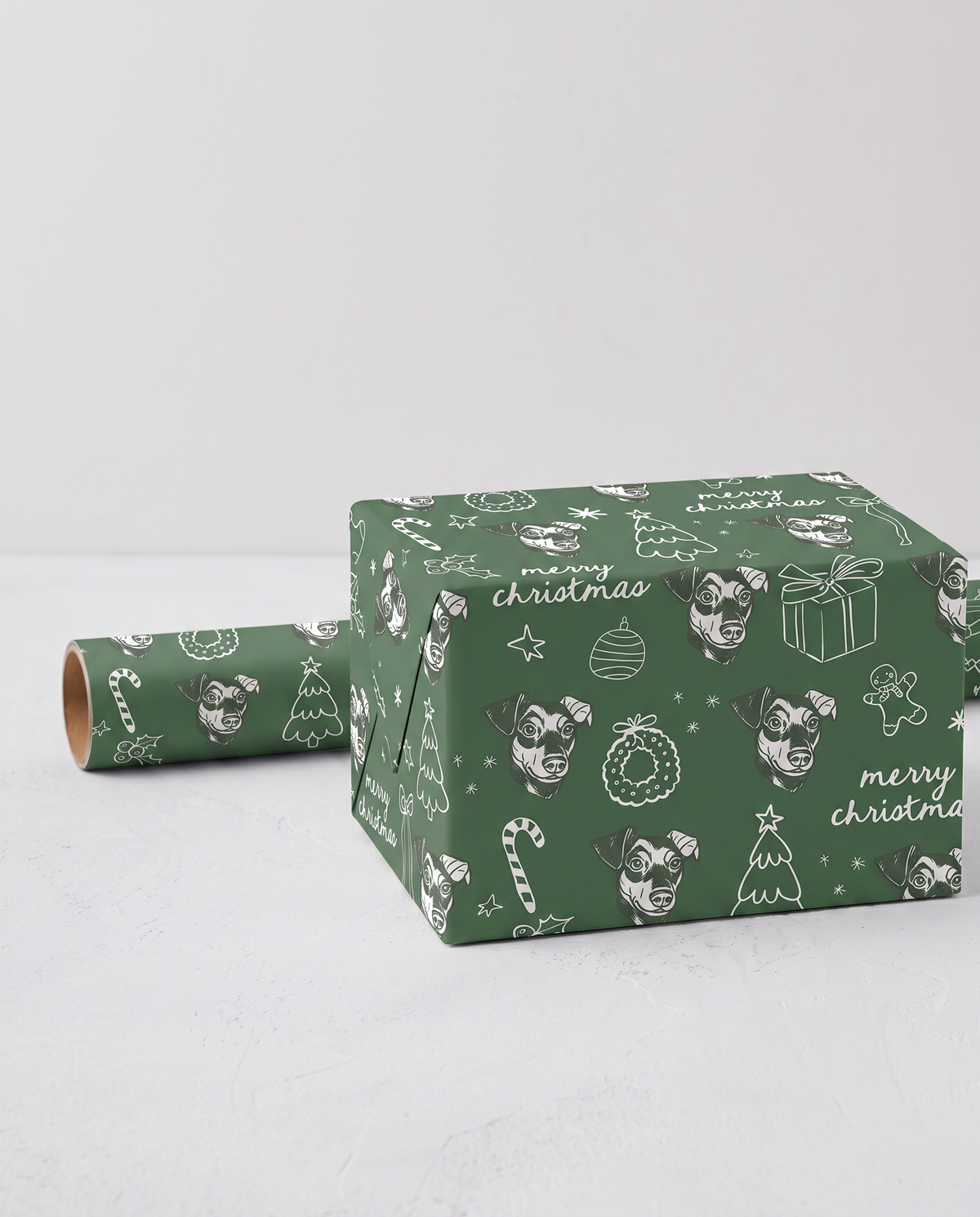 Green Christmas-themed wrapping paper with white illustrations of dog faces, candy canes, ornaments, and 'merry christmas' text, shown wrapped around a box with a matching roll beside it on a light gray background.