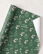 Green wrapping paper with a white line-art pattern of dogs, Christmas trees, candy canes, ornaments, and 'merry christmas' text, shown partially rolled on a light textured surface.