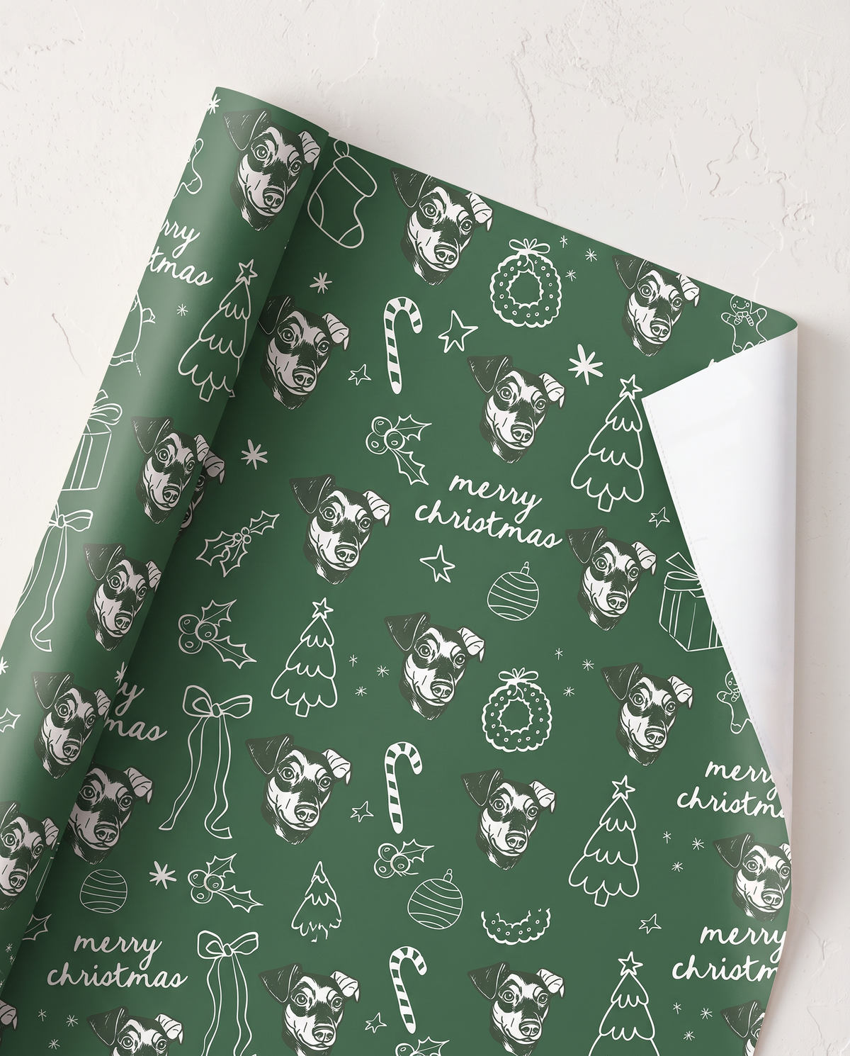 Green wrapping paper with a white line-art pattern of dogs, Christmas trees, candy canes, ornaments, and 'merry christmas' text, shown partially rolled on a light textured surface.