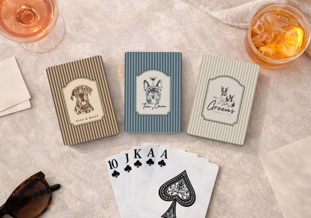 Three playing card packs with dog illustrations on a table with drinks and sunglasses.