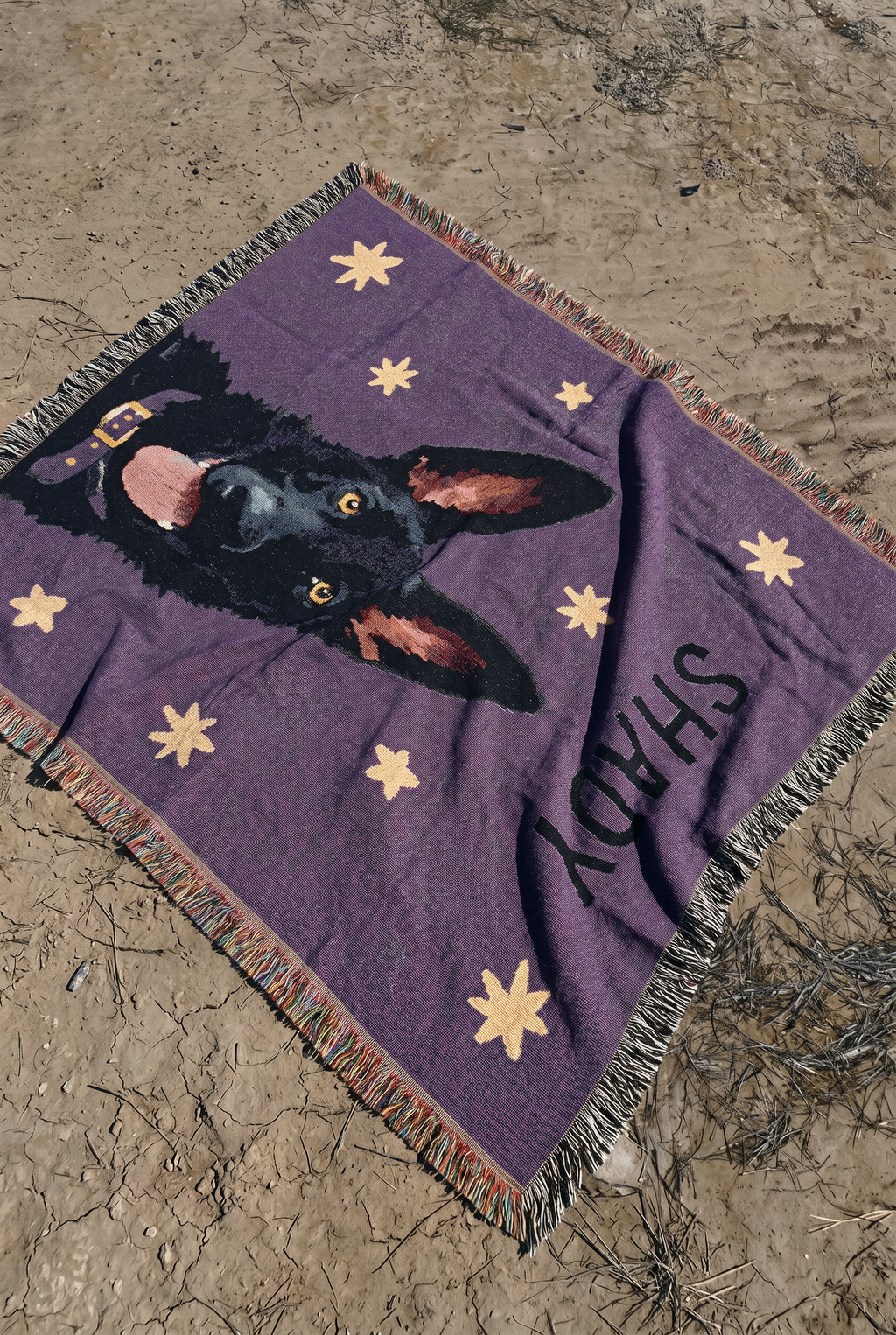 A purple woven blanket with a black dog german shepherd portrait, beige stars, and the name 'SHADY' printed in black, laid out on sandy ground.