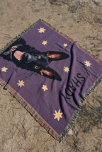 A purple woven blanket with a black dog german shepherd portrait, beige stars, and the name 'SHADY' printed in black, laid out on sandy ground.