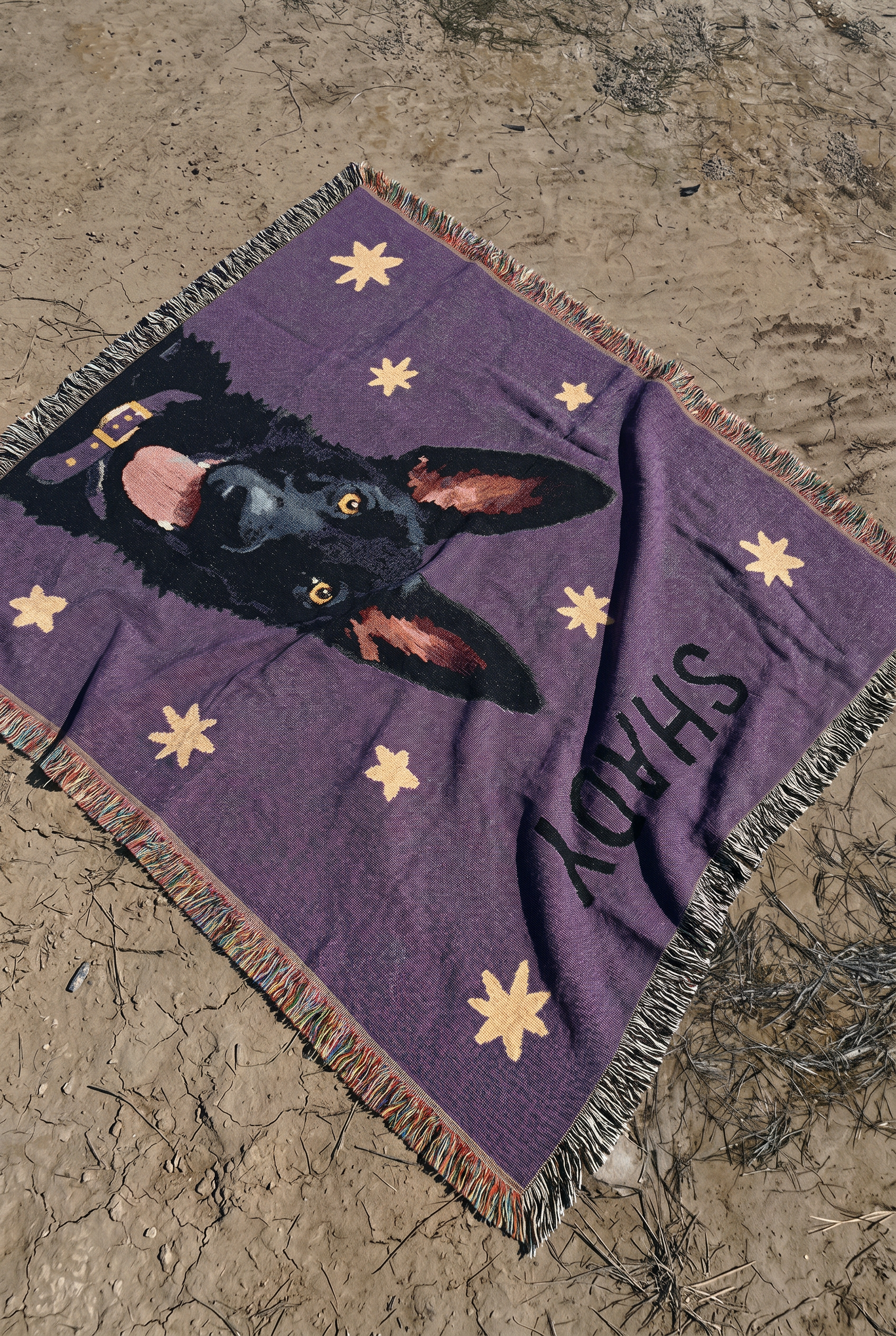 A purple woven blanket with a black dog german shepherd portrait, beige stars, and the name 'SHADY' printed in black, laid out on sandy ground.