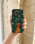 A hand with dark green nail polish holds a dark green phone case featuring a brown dachshund illustration, white star details, and the name 'dixie' in white script, set against a light beige stone wall background.