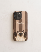 A personalized phone case featuring a brown and beige striped background, a Siamese cat illustration with raised paws, and the name 'Caroline' in white script. The case is shown on a light-colored, textured surface.