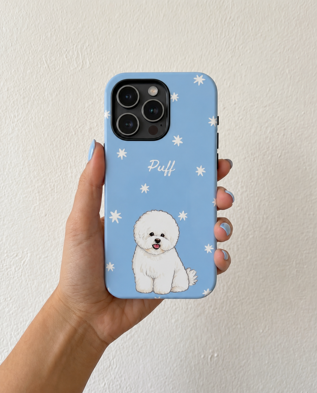A hand holding a light blue phone case featuring a white cartoon dog illustration and the name 'Puff' in white script, set against a plain off-white wall.