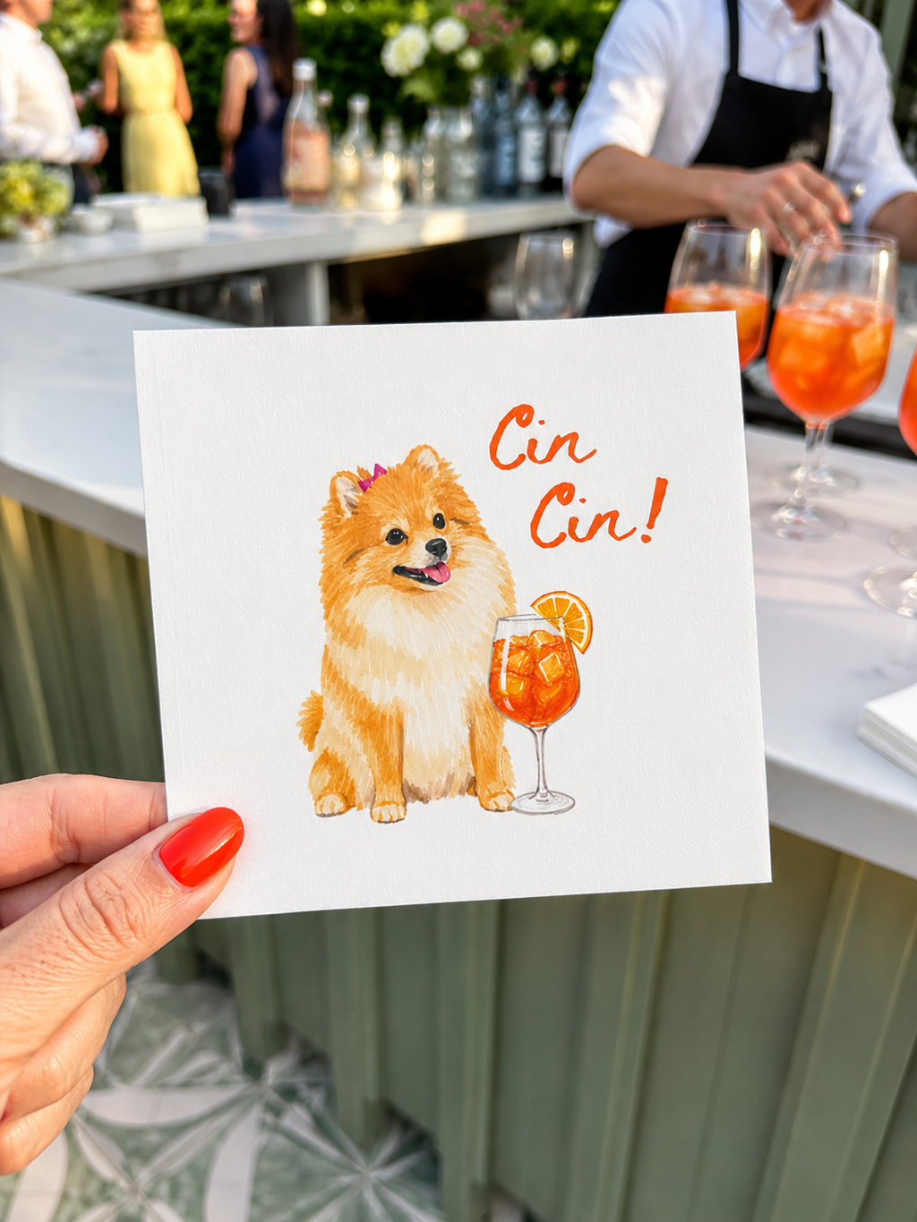 A hand with red nail polish holds a square white greeting card featuring a watercolor illustration of a fluffy golden dog next to an orange cocktail with the text 'Cin Cin!'. The background shows a blurred outdoor bar or patio scene with people and drinks.