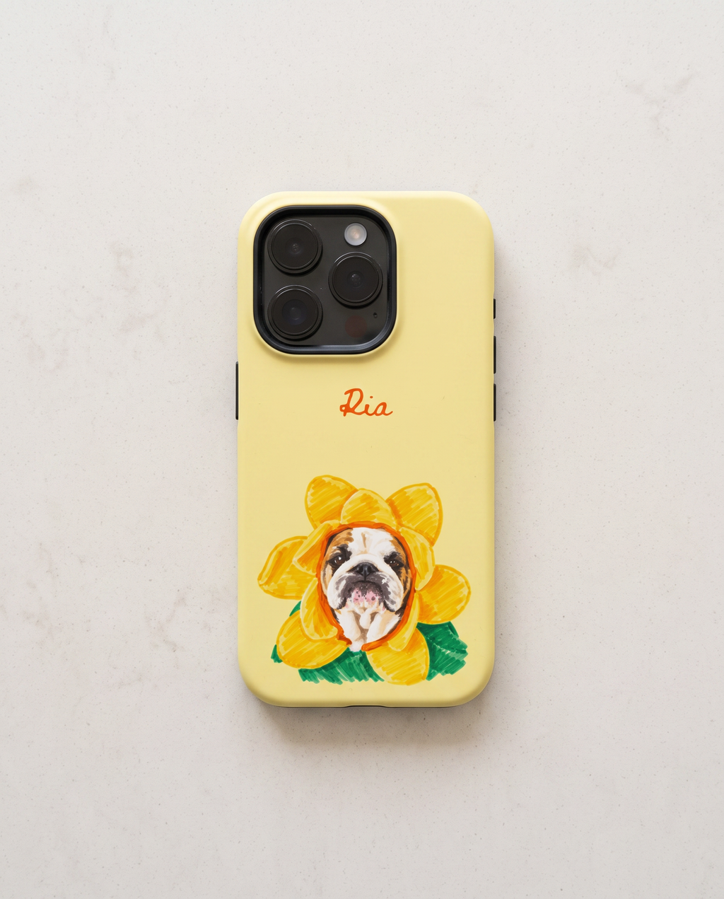 A pale yellow phone case featuring a custom illustration of a bulldog inside a yellow flower, with the name 'Ria' written in red cursive above it. The case is shown on a light beige marble surface.
