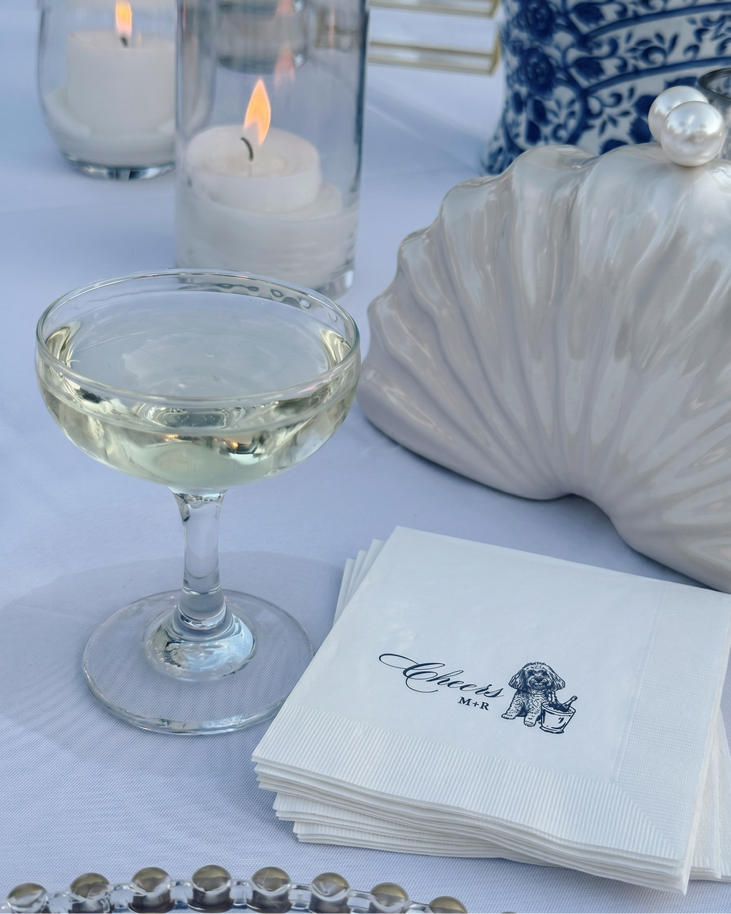 Cocktail glass with a drink, stack of napkins, and decorative shell on a tablecloth.