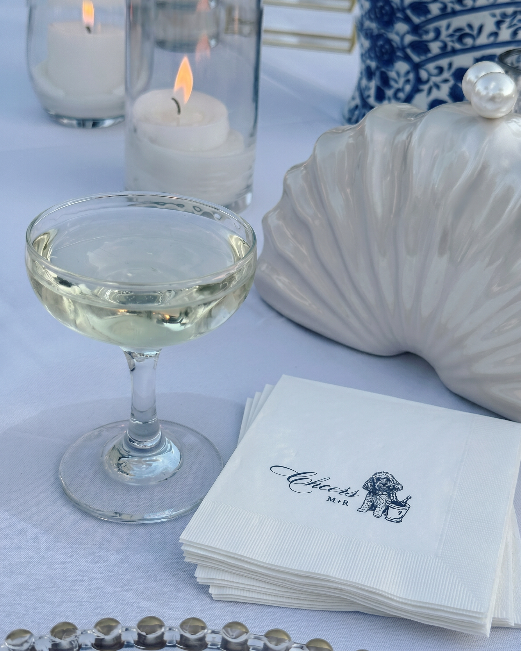 Cocktail glass with a drink, stack of napkins, and decorative shell on a tablecloth.