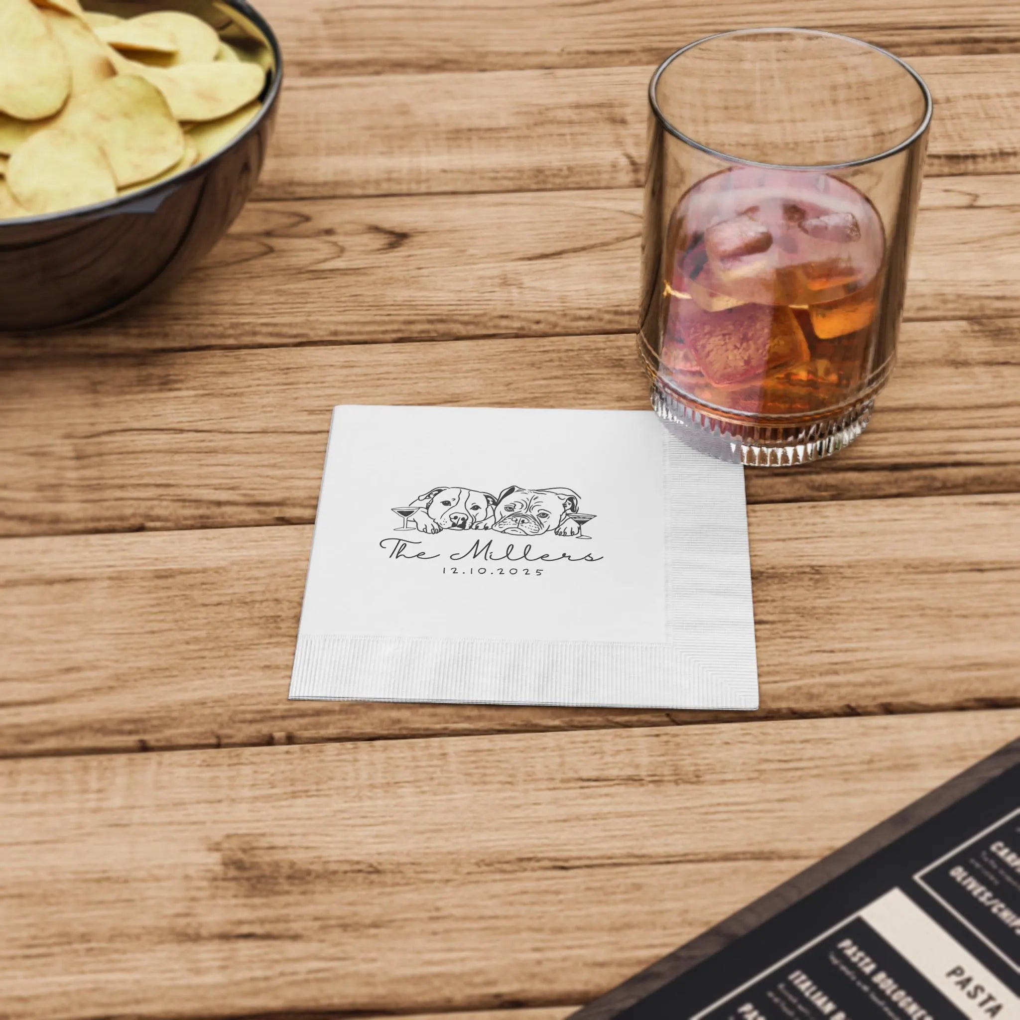 Glass of whiskey with ice and a bowl of chips on a wooden table, featuring dog napkin.