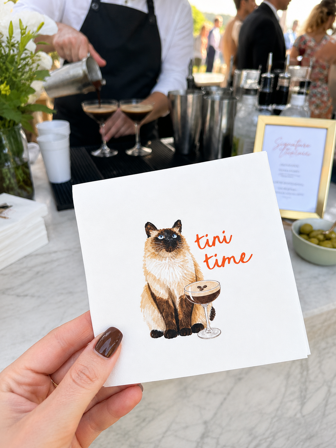 A hand holds a white square card featuring an illustration of a Siamese cat next to a cocktail glass with the text 'tini time' in orange. The background shows a blurred outdoor bar scene with a bartender pouring drinks and people.