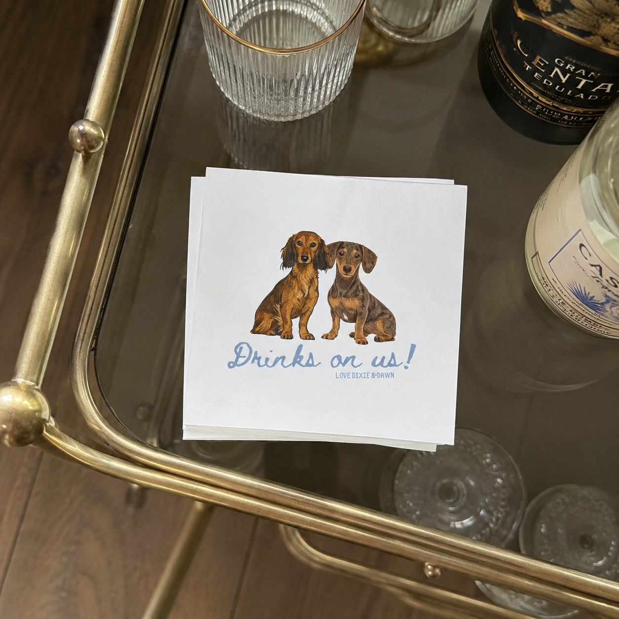 A stack of white paper napkins featuring an illustration of two dachshunds and the text 'Drinks on us!' h, placed on a gold-framed glass table with barware and bottles around it.