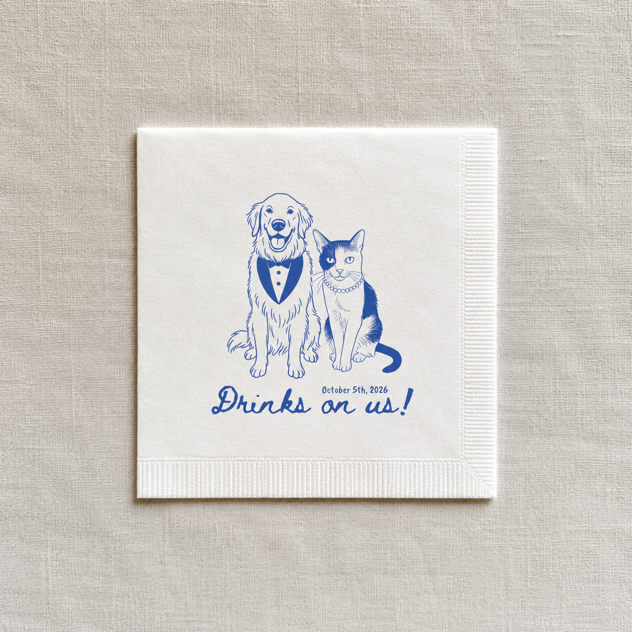 A square white paper napkin with blue line art of a golden retriever and a cat wearing a bow tie, with the text 'Drinks on us!' and a date printed below, displayed on a textured beige linen background.
