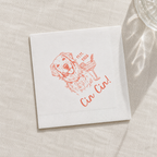 A square white paper napkin with a red line-art illustration of a dog holding a cocktail glass and the text 'Cin Cin!' and '11th 2026', placed on a light beige textured surface with soft shadows.