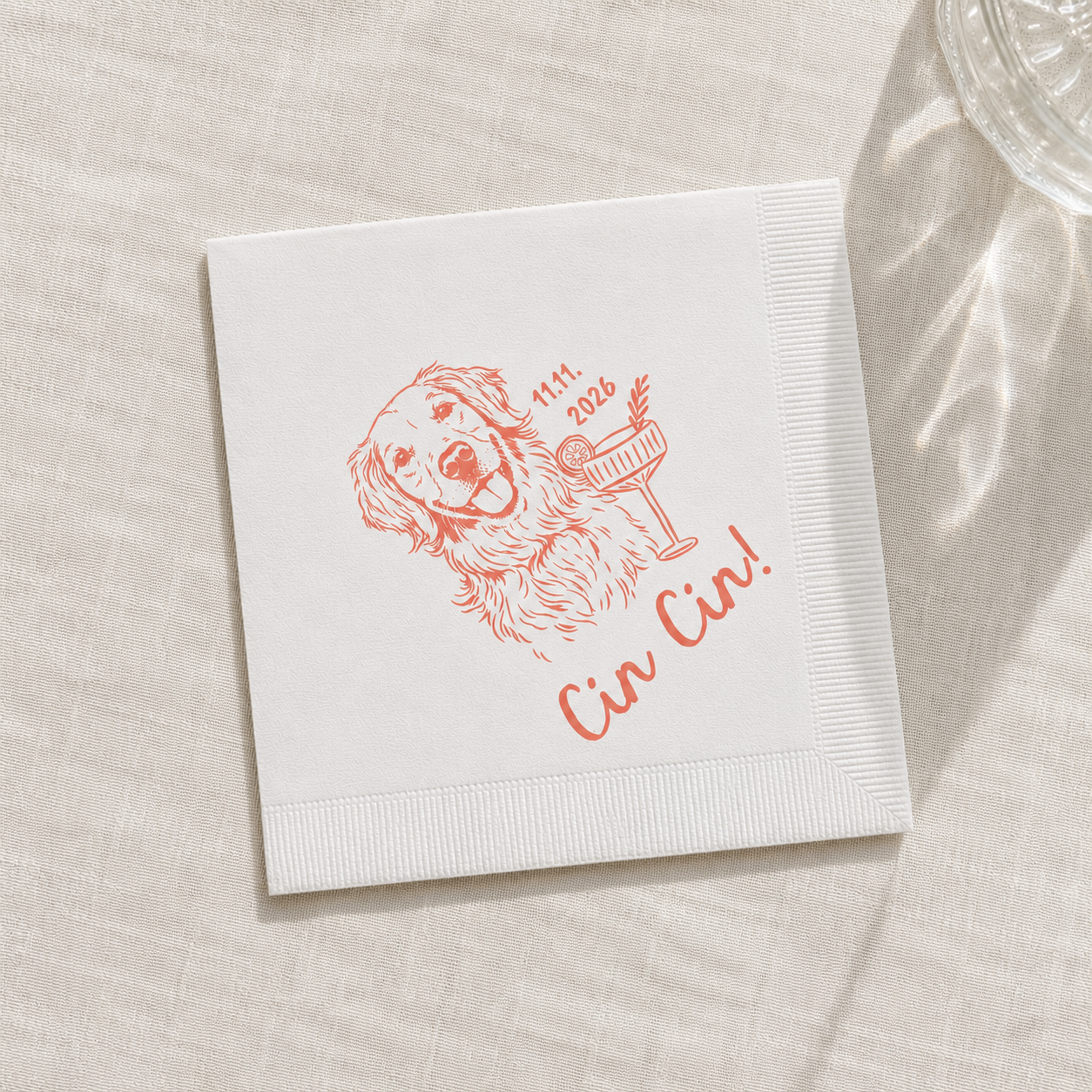 A square white paper napkin with a red line-art illustration of a dog holding a cocktail glass and the text 'Cin Cin!' and '11th 2026', placed on a light beige textured surface with soft shadows.