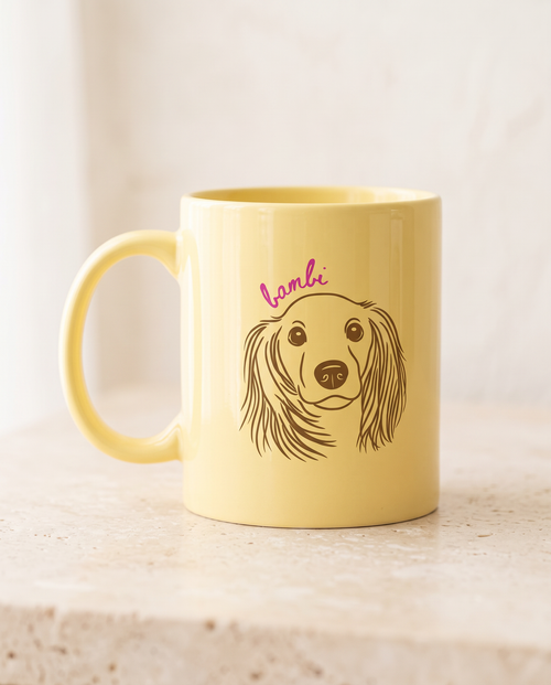 A pale yellow ceramic mug with a brown line art illustration of a long-eared dog and the word 'lambi' in pink script above it, shown on a light stone surface against a soft off-white background.
