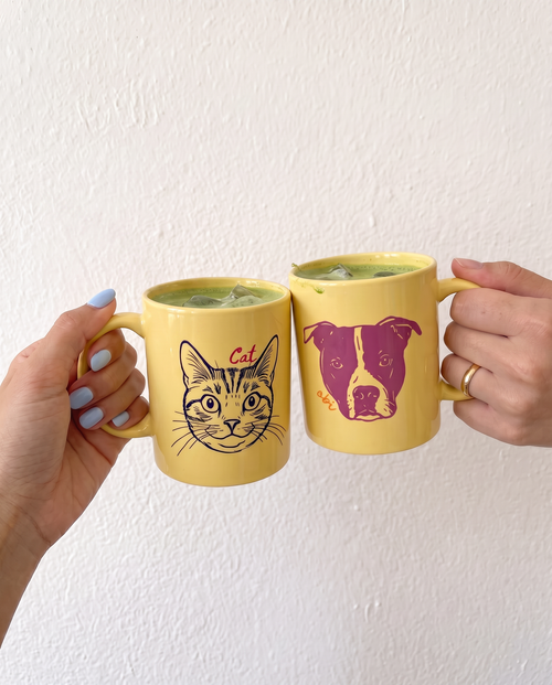 Two yellow ceramic mugs held by hands against a white textured wall. One mug features a black line art cat illustration with the word 'Cat' in red, and the other features a purple dog face illustration.
