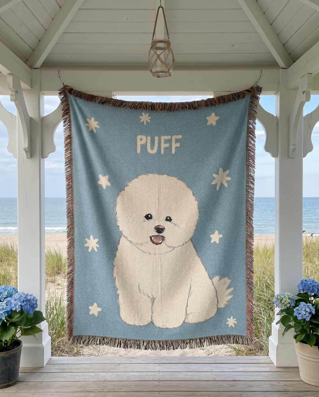 A woven beach-themed tapestry featuring a white Bichon Frise dog named 'PUFF' on a light blue background with cream stars, hanging on a white porch with an ocean view in the background.