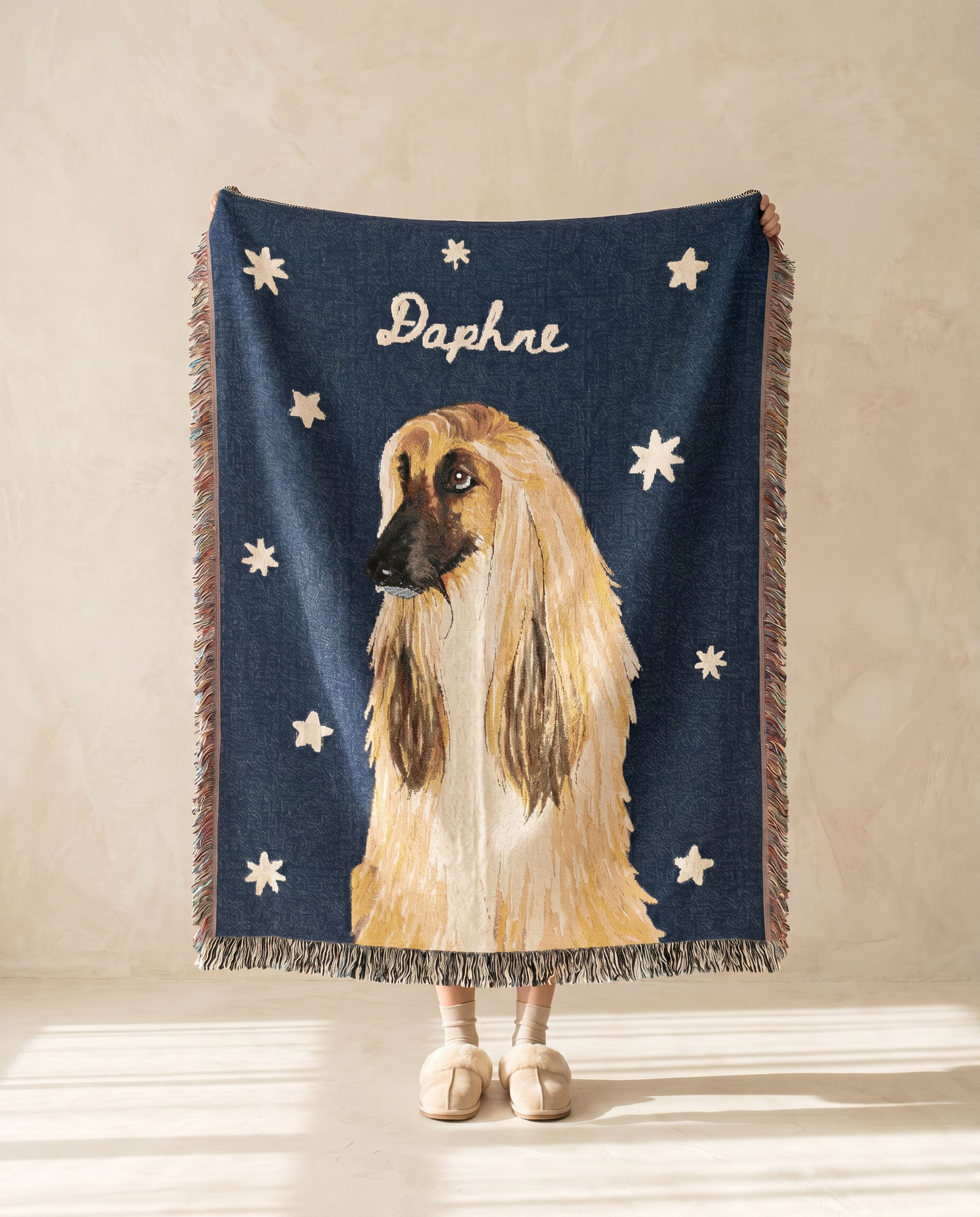 A navy blue woven throw blanket with fringe, featuring a large illustration of a beige and white Afghan hound, white stars, and the name 'Daphne' in white script. The blanket is held up by a person whose legs and feet are visible below, standing against a warm beige textured wall.