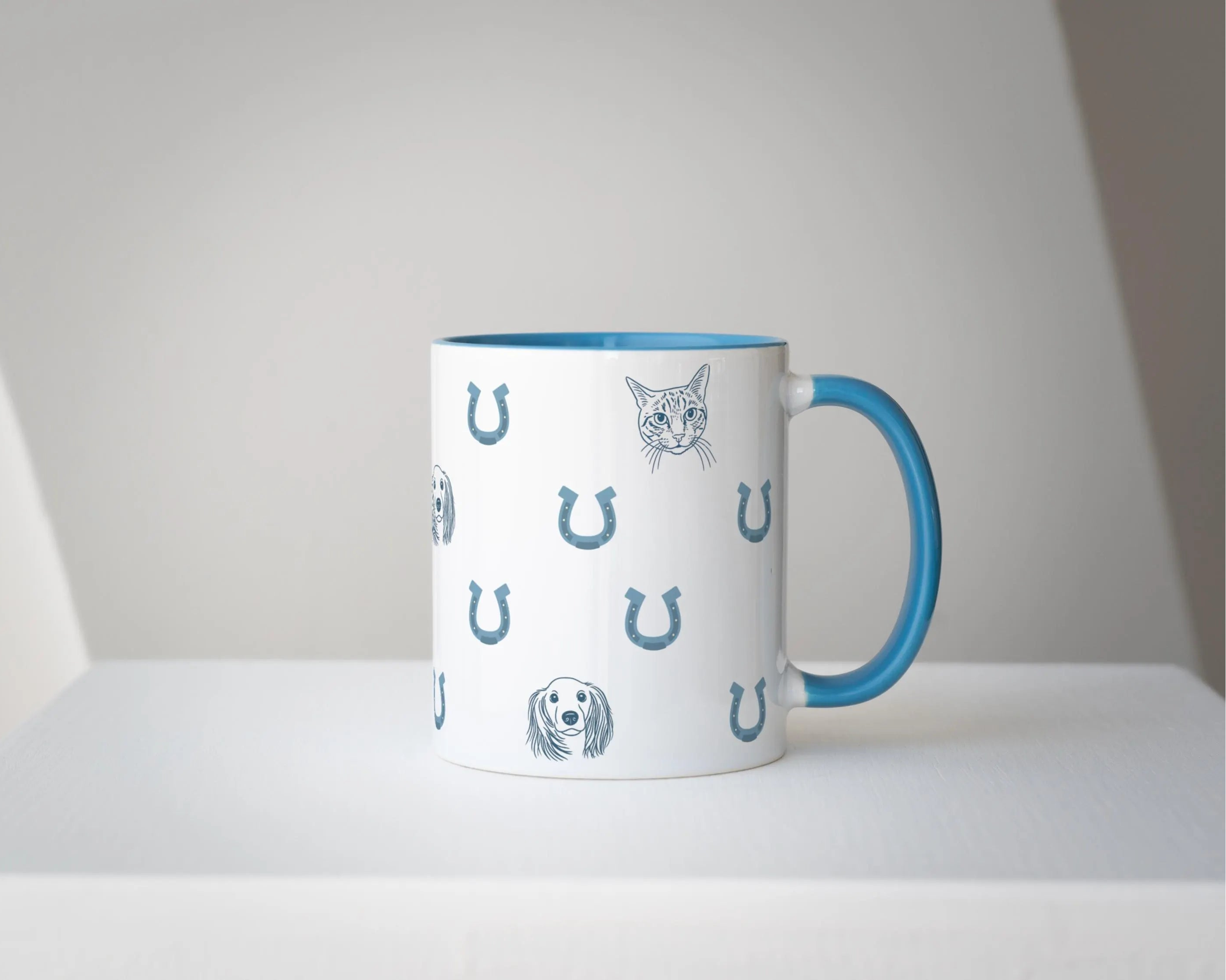 Custom mug with cat and dog illustrations and horseshoes and blue handle on a white surface