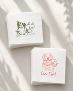 Two stacks of white cocktail napkins on a light beige surface with soft leaf shadows. One stack has a green dog illustration and the text 'I do too!' with 'LOVE, MAX' below.