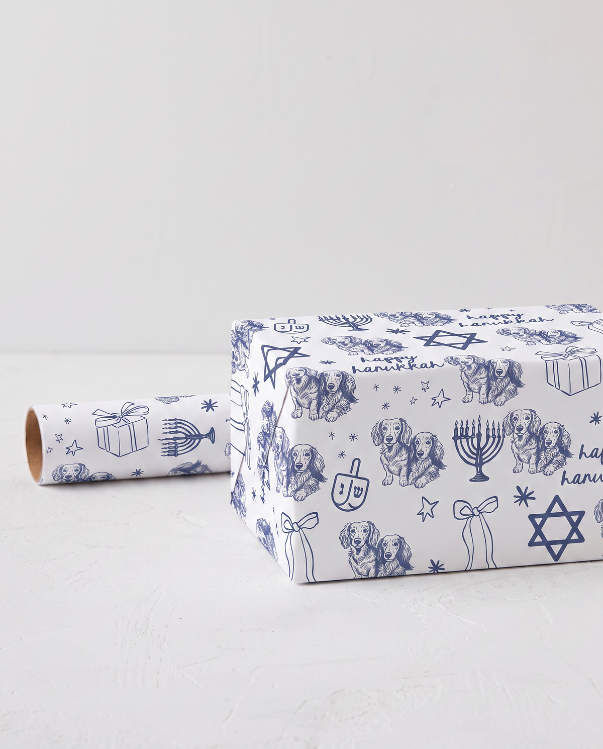 Dachshund dog doodles on White wrapping paper with a navy blue Hanukkah-themed pattern, shown wrapped around a rectangular gift box with a matching roll of paper on a light gray surface.