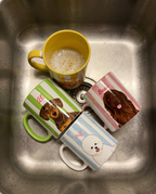 Four dog-themed ceramic mugs with different stripe patterns and dog illustrations are arranged in a stainless steel kitchen sink.