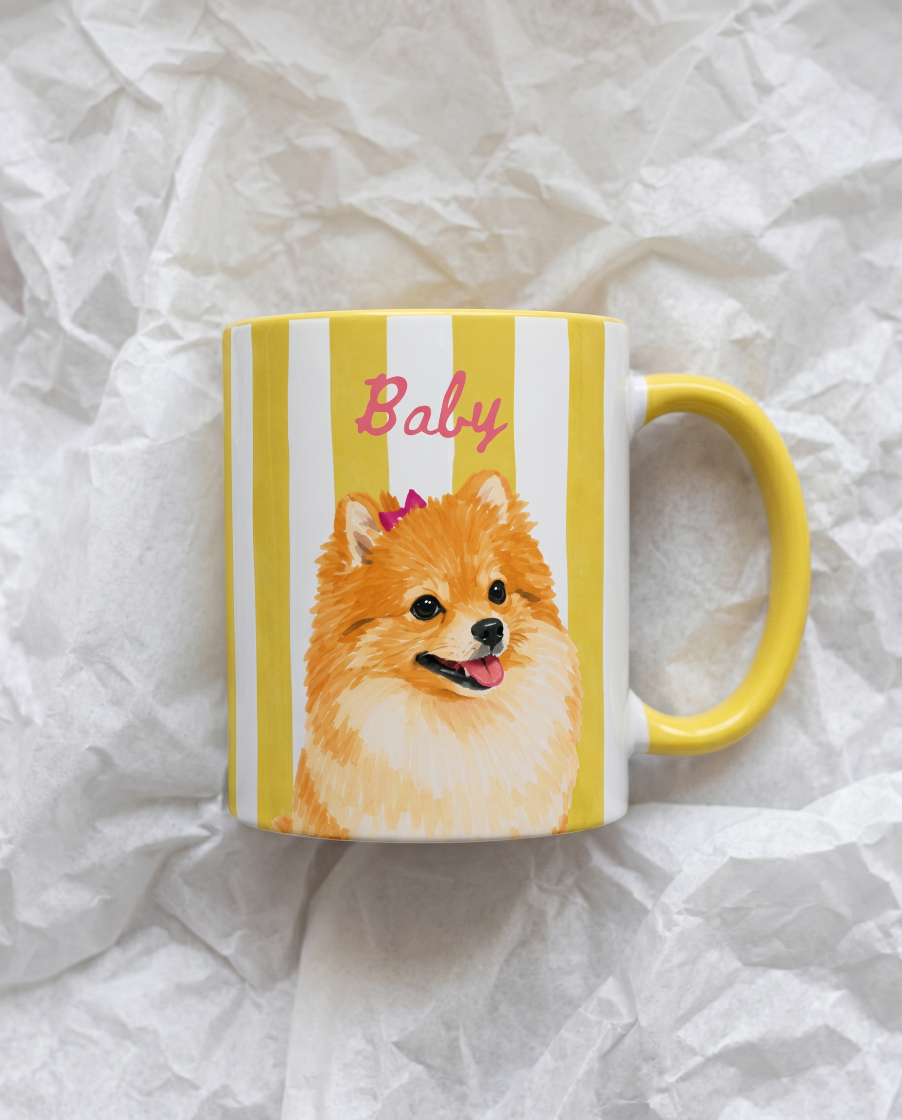 A white ceramic mug with yellow and white vertical stripes, featuring a printed illustration of a fluffy golden Pomeranian dog wearing a pink crown and the word 'Baby' in pink script. The mug has a solid yellow handle and is photographed on crumpled white paper.