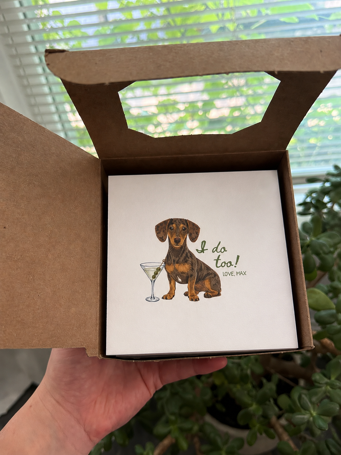 A hand holds an open brown cardboard gift box containing a square white card with an illustration of a dachshund sitting next to a martini glass and the text 'I do too! LOVE, MAX'. The background shows a window with blinds and green plants.