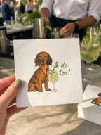 A hand holds a white square card featuring a watercolor illustration of a brown dachshund sitting next to a green cocktail glass with the text 'I do too!' in green script. The background shows an outdoor event or bar setting with people and drinks blurred.