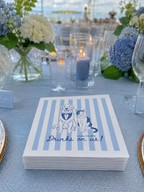A stack of white paper napkins with light blue vertical stripes and a navy blue illustration of a dog and cat wearing tuxedos, with the text 'Drinks on us!' printed below. The napkins are displayed on a light blue tablecloth at an outdoor event table with flowers, candles, and glassware in the background.