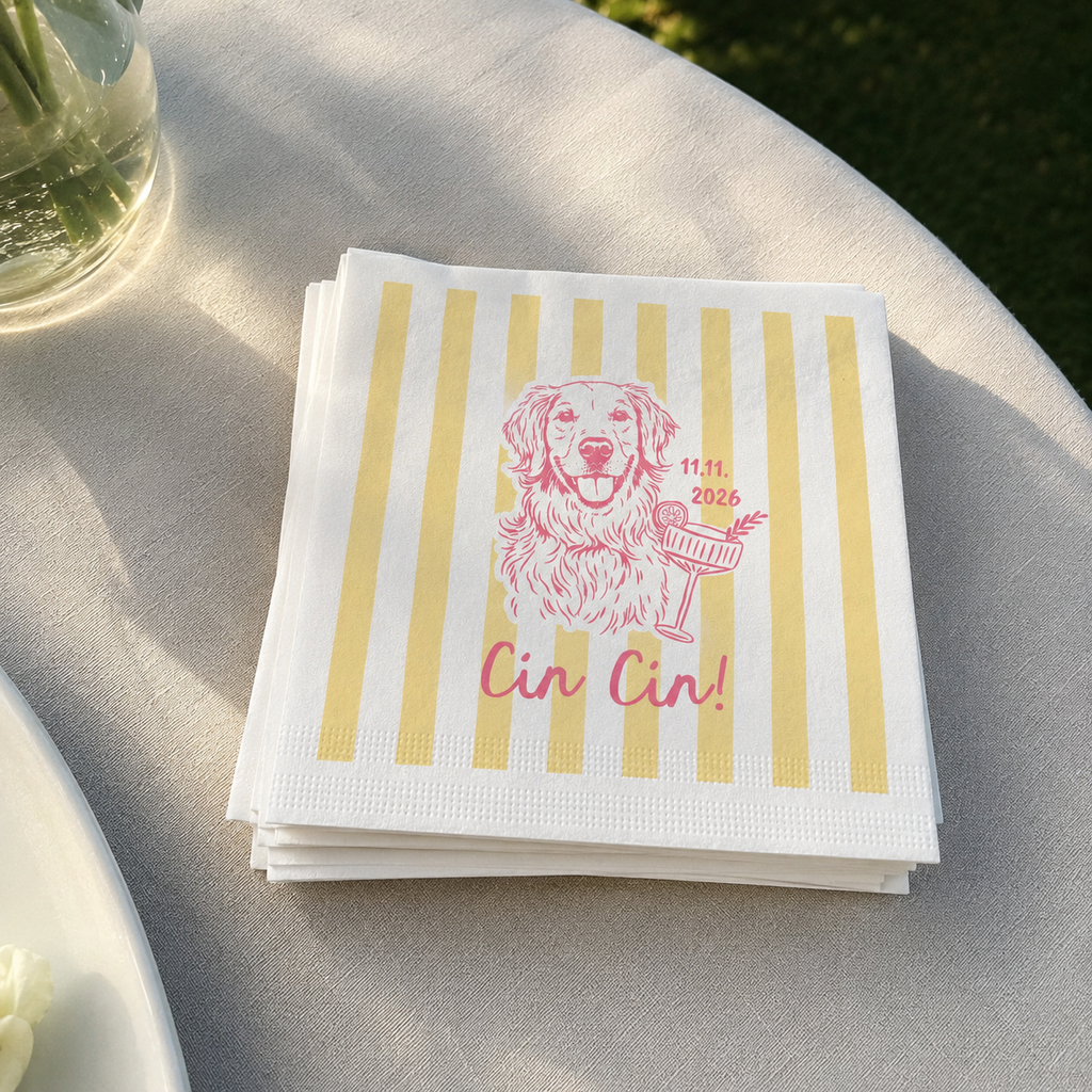 A stack of white paper napkins with vertical yellow stripes and a pink line drawing of a dog holding a cocktail, with the text 'Cin Cin!' printed below. The napkins are placed on a light-colored tablecloth outdoors.
