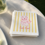 A stack of white paper napkins with vertical yellow stripes and a pink line drawing of a dog holding a cocktail, with the text 'Cin Cin!' printed below. The napkins are placed on a light-colored tablecloth outdoors.