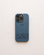 A slate blue phone case with a black engraved design of two puppies at the bottom and the initials 'B.E.