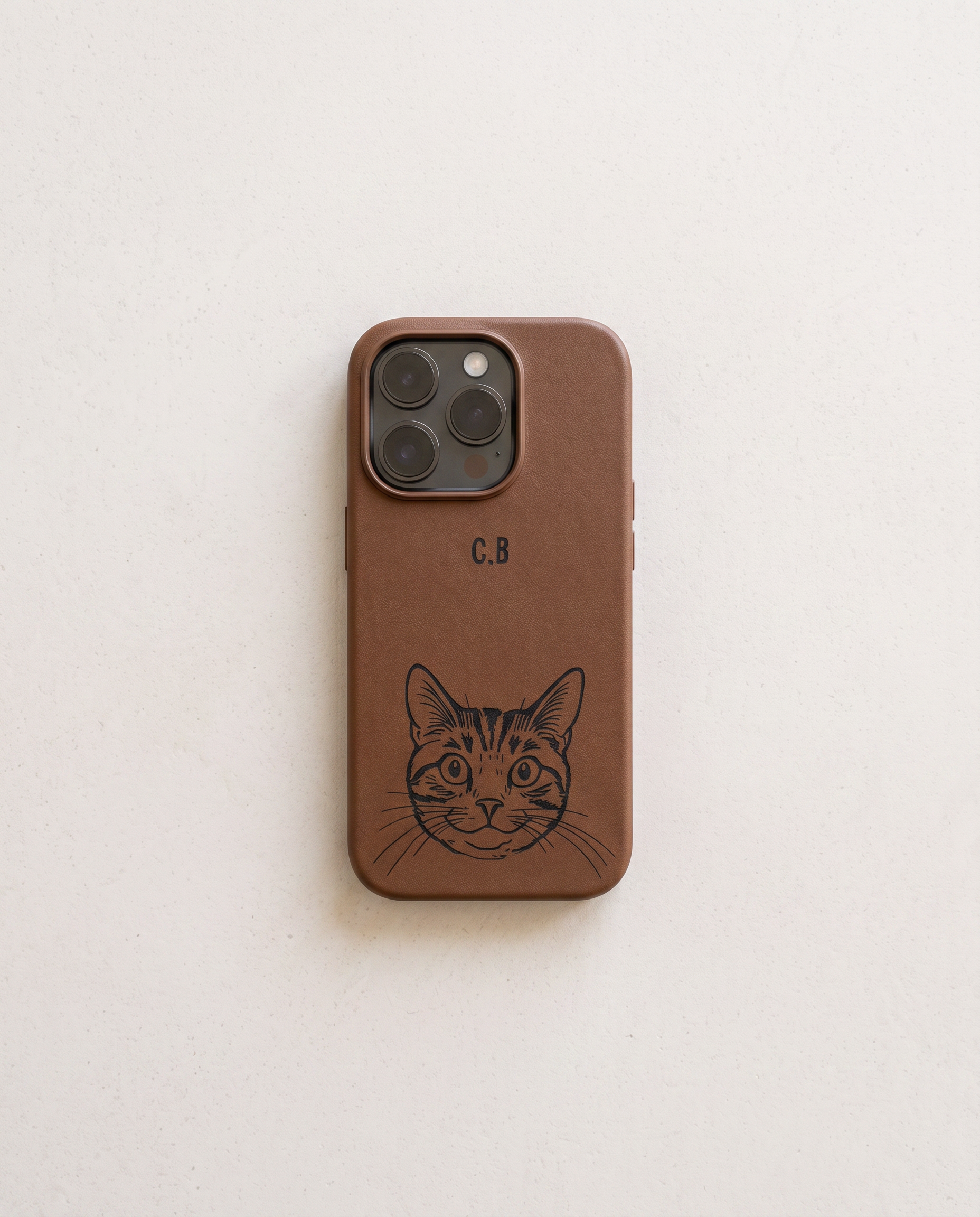 A brown leather phone case with a black engraved cat face illustration and the initials 'C.B' on the back, shown on a light off-white background.