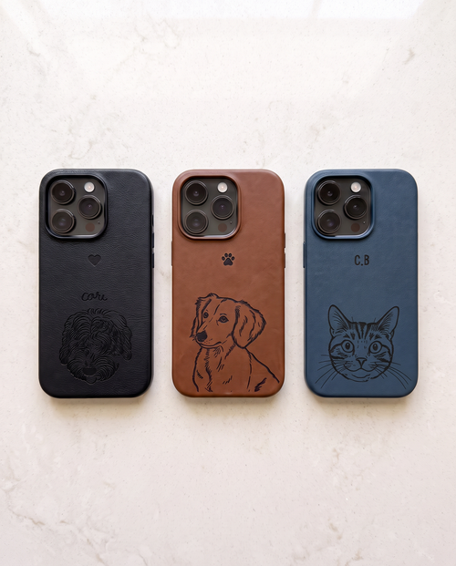 Three custom leather phone cases with camera cutouts are displayed side-by-side on a light marble surface. The left case is black with an embossed dog head and the name 'C.