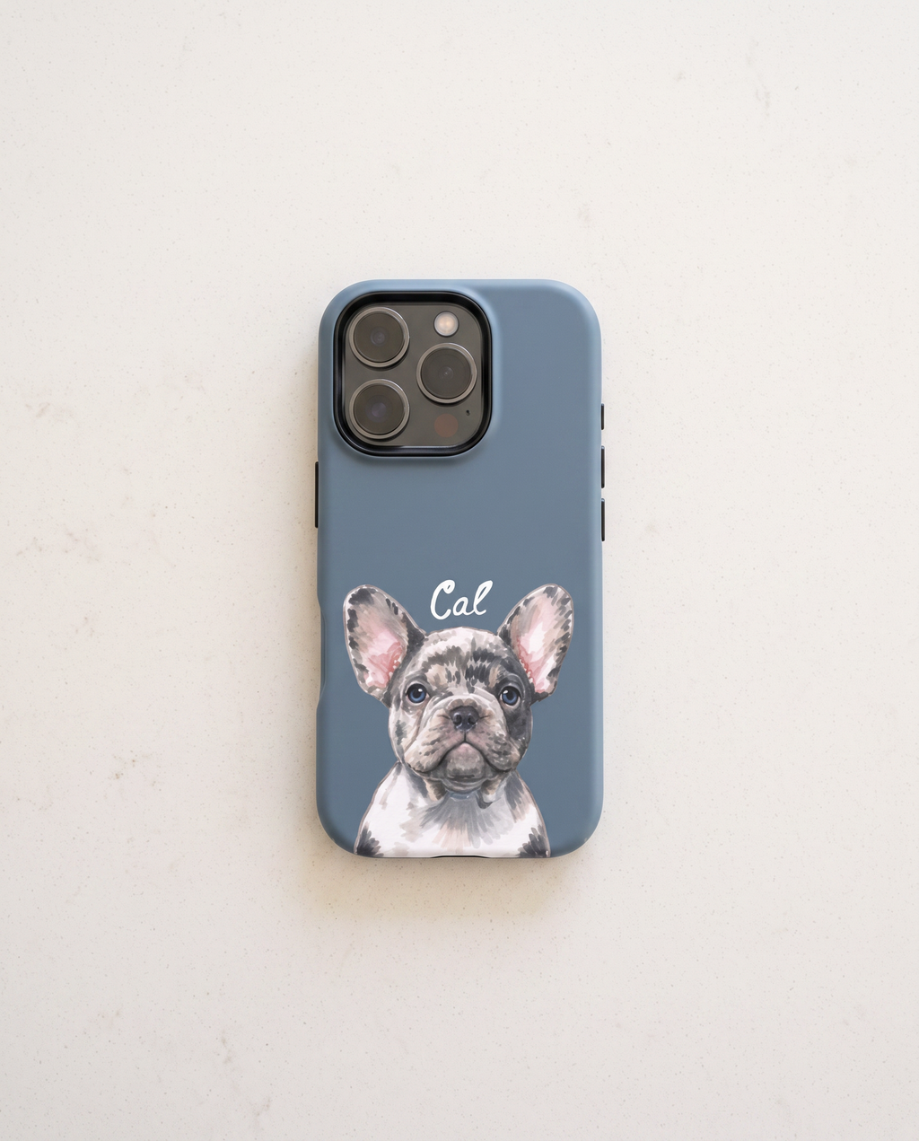 A slate blue phone case featuring a printed illustration of a French Bulldog named 'Cal' in white script, displayed on a light beige textured surface.