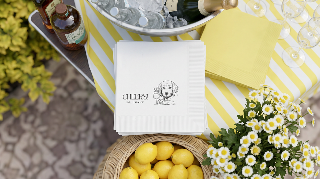 Outdoor bar set up with dog cocktail napkins, lemons, flowers, and bottles on a striped tablecloth