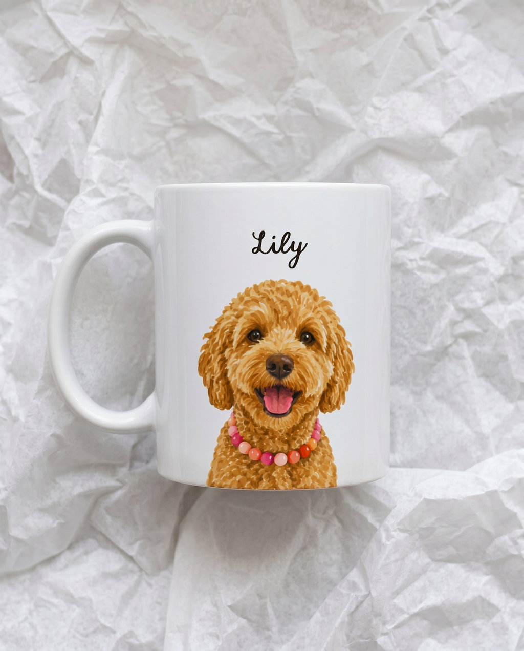 A white ceramic mug featuring a printed illustration of a smiling golden doodle dog wearing a pink beaded collar, with the name 'Lily' written above in black script. The mug is photographed on crumpled white paper.