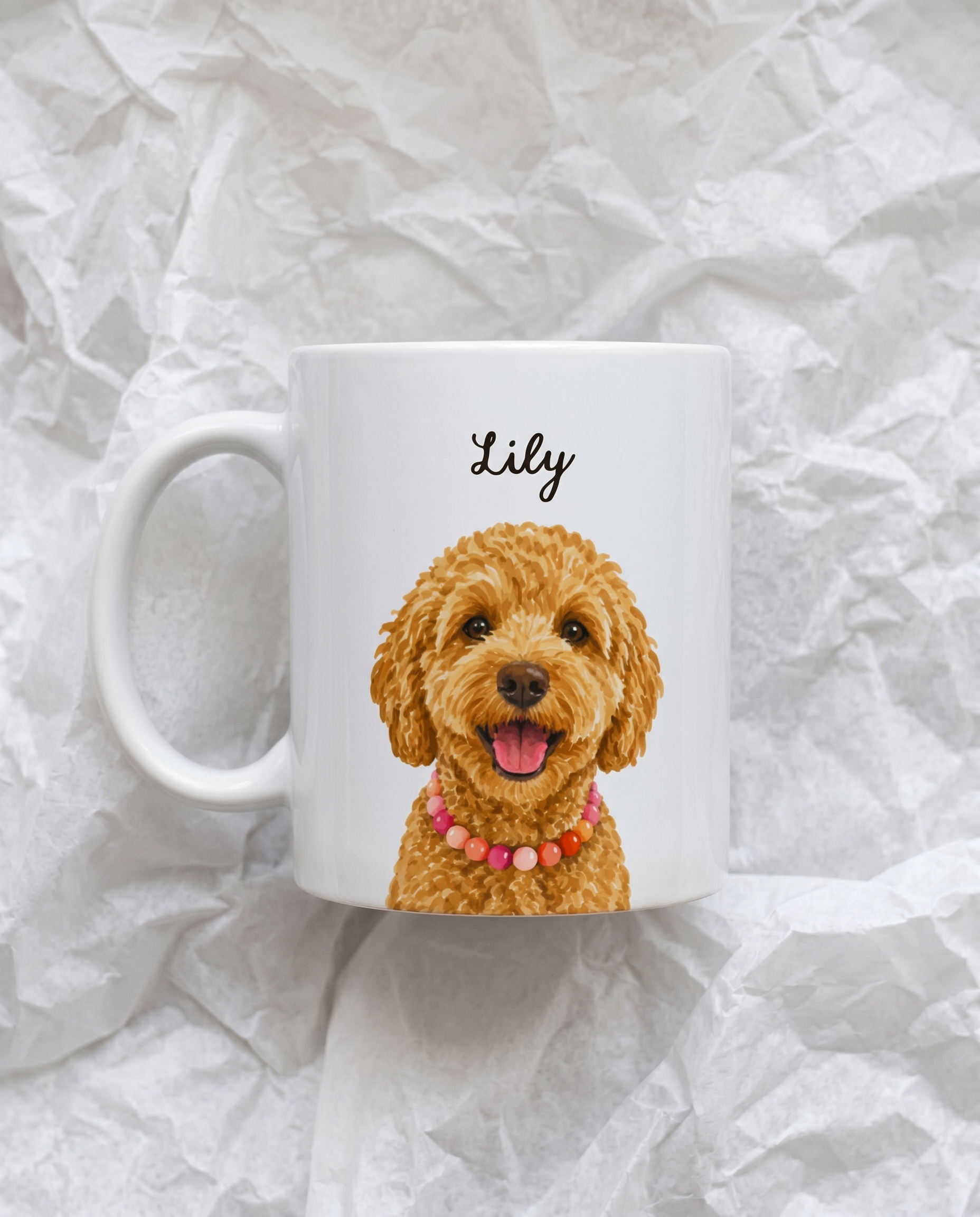 A white ceramic mug featuring a printed illustration of a smiling golden doodle dog wearing a pink beaded collar, with the name 'Lily' written above in black script. The mug is photographed on crumpled white paper.