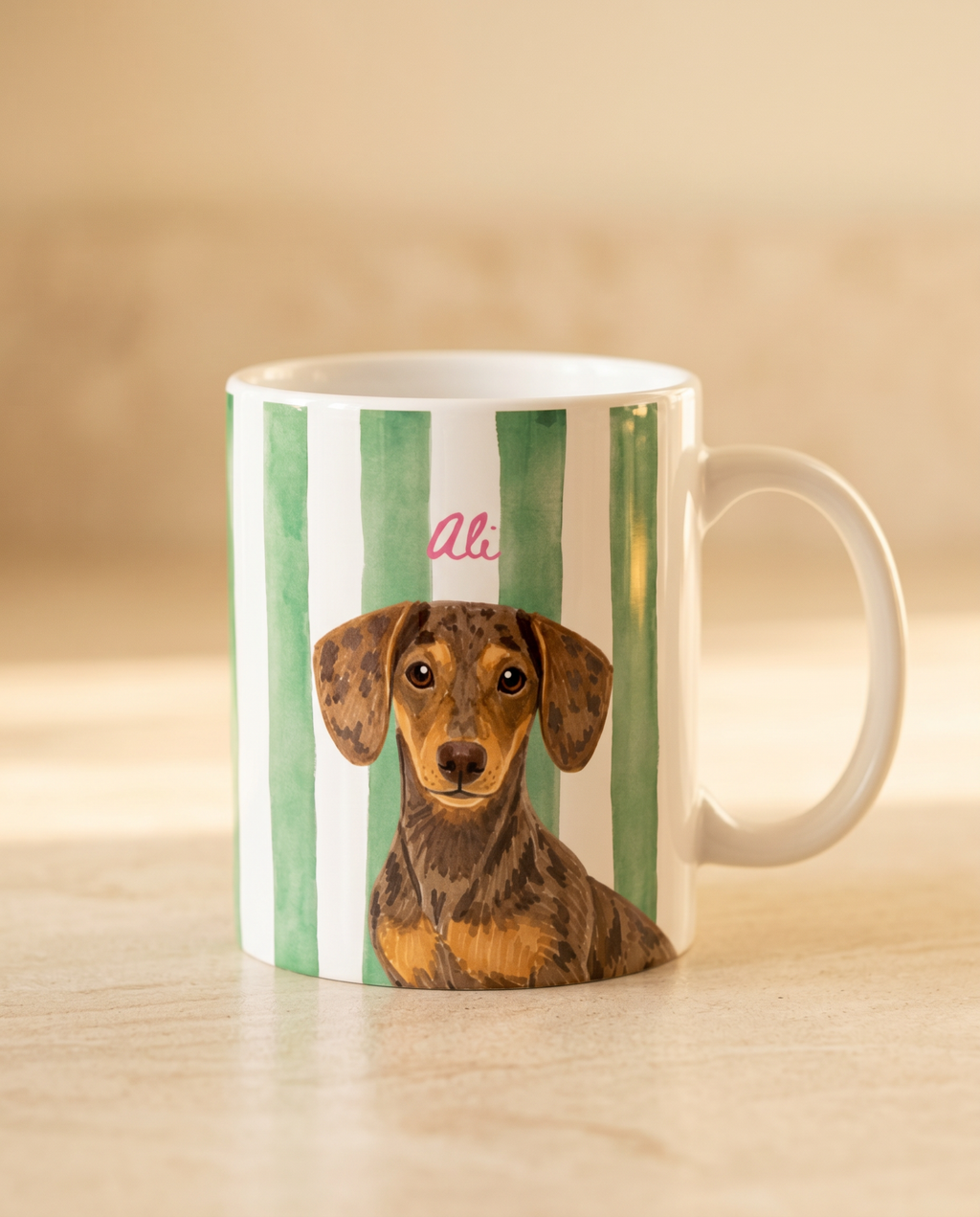 A white ceramic mug with vertical green stripes and a printed portrait of a dachshund, featuring the pink cursive name 'Ali' above the dog, set on a light wooden surface.