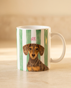 A white ceramic mug with vertical green stripes and a printed portrait of a dachshund, featuring the pink cursive name 'Ali' above the dog, set on a light wooden surface.
