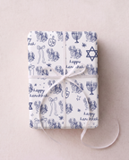 A wrapped gift box tied with a white ribbon, covered in white and blue hannukah wrapping paper printed with navy blue illustrations of dogs, dreidels, Star of David, menorahs, and the words 'happy hanukkah'.