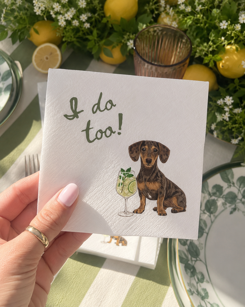 A hand holding a white textured paper napkin printed with the phrase 'I do too!' in green script and an illustration of a dachshund sitting next to a cocktail glass. The napkin is held over a styled table setting with lemons, greenery, and plates.
