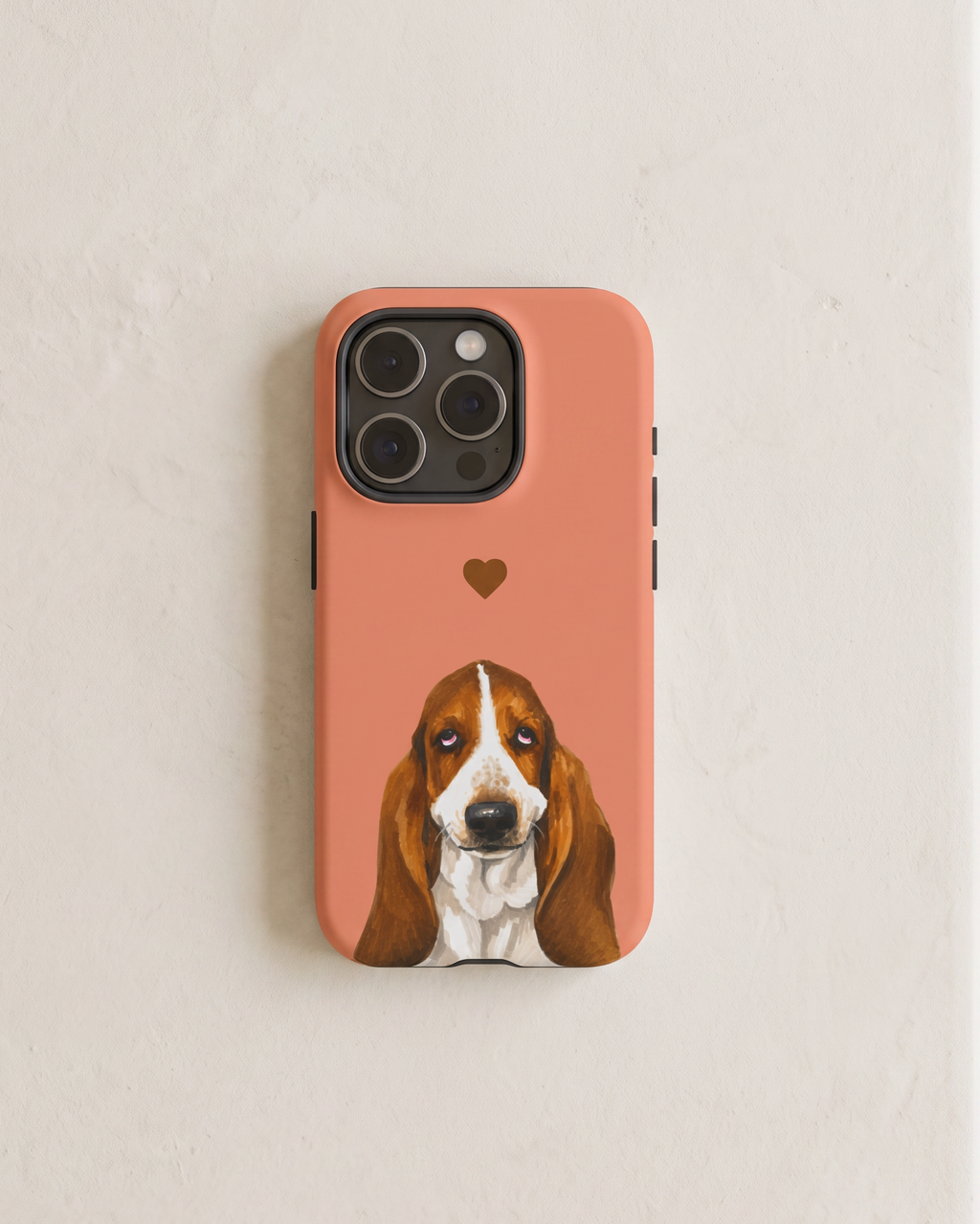 A coral pink phone case featuring a printed illustration of a Basset Hound dog and a small heart, shown on a light beige textured wall.